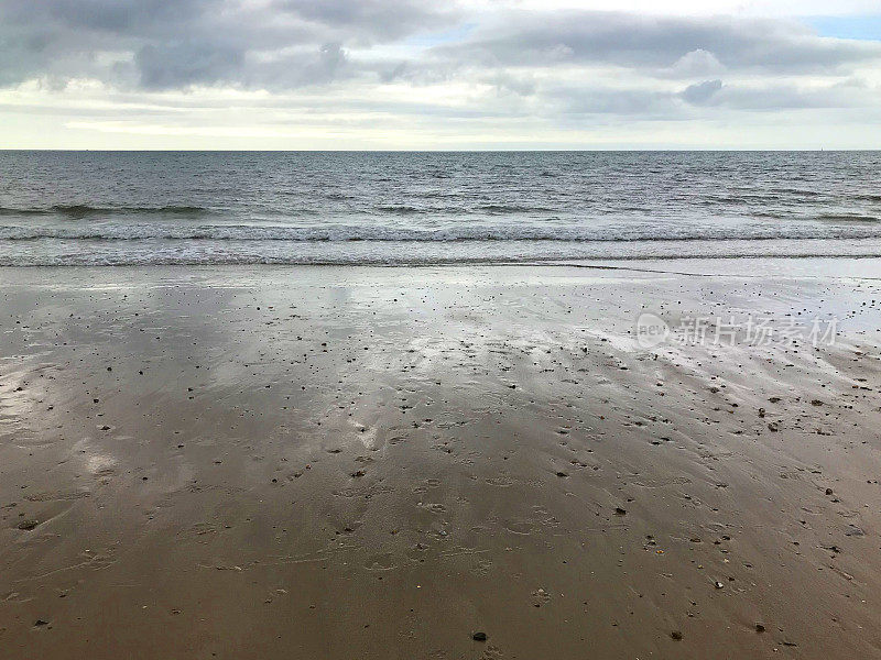 暴风雨的海滩日清晨，灰色的风暴云聚集在头顶上，黑暗的大海，温和的海浪，潮水拍打着海边的海岸线和海滩边潮湿的沙子，文字复制空间，沙滩上的脚印/狗的脚印，英国